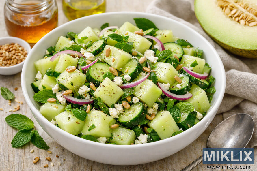 A bright, fresh honeydew cucumber salad served in a white ceramic bowl on a wooden surface, garnished with feta cheese, red onion, mint, and toasted seeds. A bright, fresh honeydew cucumber salad served in a white ceramic bowl on a wooden surface, garnished with feta cheese, red onion, mint, and toasted seeds.