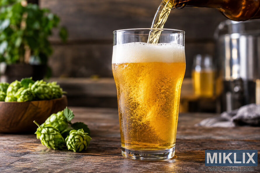 Fresh golden IPA pouring from an unbranded brown bottle into a clear pint glass with a creamy foam head, surrounded by green hops on a rustic wooden table. Fresh golden IPA pouring from an unbranded brown bottle into a clear pint glass with a creamy foam head, surrounded by green hops on a rustic wooden table.