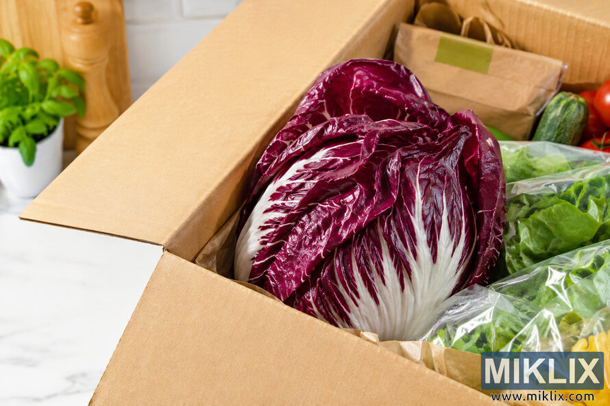 Fresh purple radicchio displayed inside an unbranded cardboard grocery delivery box alongside leafy greens and fresh vegetables on a bright kitchen countertop with soft natural daylight. Fresh purple radicchio displayed inside an unbranded cardboard grocery delivery box alongside leafy greens and fresh vegetables on a bright kitchen countertop with soft natural daylight.