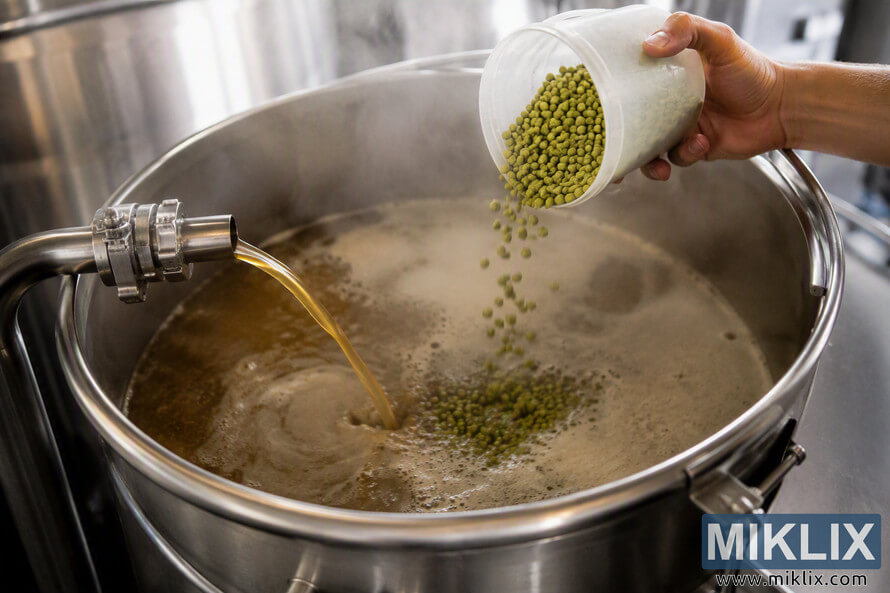 Close-up landscape view of a brewing process showing green hop pellets being poured into a large stainless steel brew kettle while amber-colored wort flows from a metal outlet during runoff. The kettle surface contains foam and swirling liquid, and the background shows blurred brewery equipment.