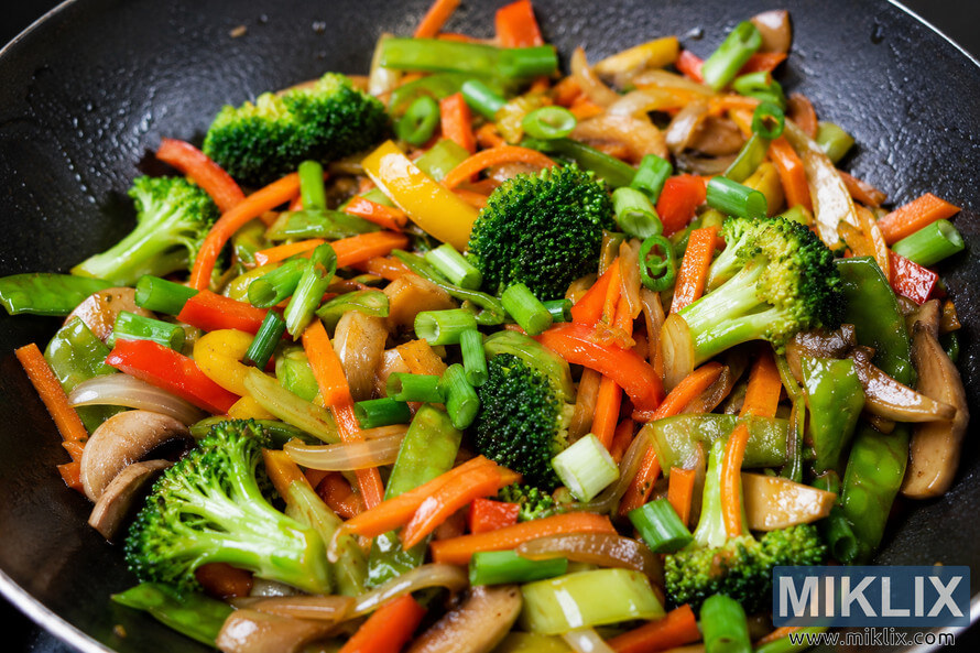 Colorful Asian vegetable stir-fry with broccoli, carrots, bell peppers, mushrooms, onions, snap peas, and chopped scallions cooking in a dark wok with a glossy sauce.