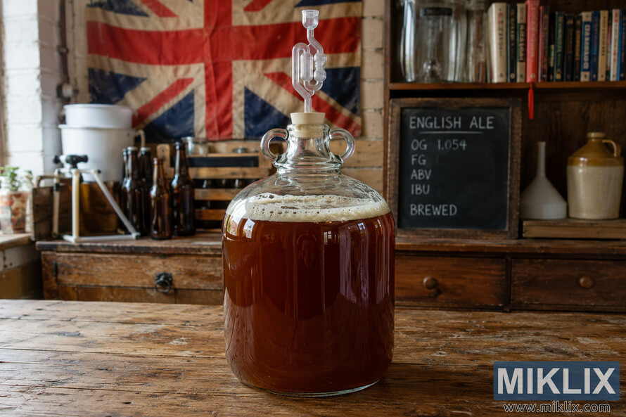 Glass carboy filled with fermenting English ale on a rustic wooden table inside a traditional British-style homebrewing workshop with vintage brewing tools and warm ambient lighting. Glass carboy filled with fermenting English ale on a rustic wooden table inside a traditional British-style homebrewing workshop with vintage brewing tools and warm ambient lighting.
