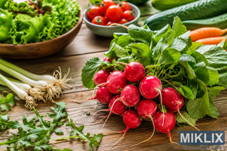 Fresh red radishes with leafy greens arranged on a rustic wooden table alongside lettuce, cherry tomatoes, cucumbers, carrots, spring onions, and arugula in natural daylight, representing a healthy lifestyle.