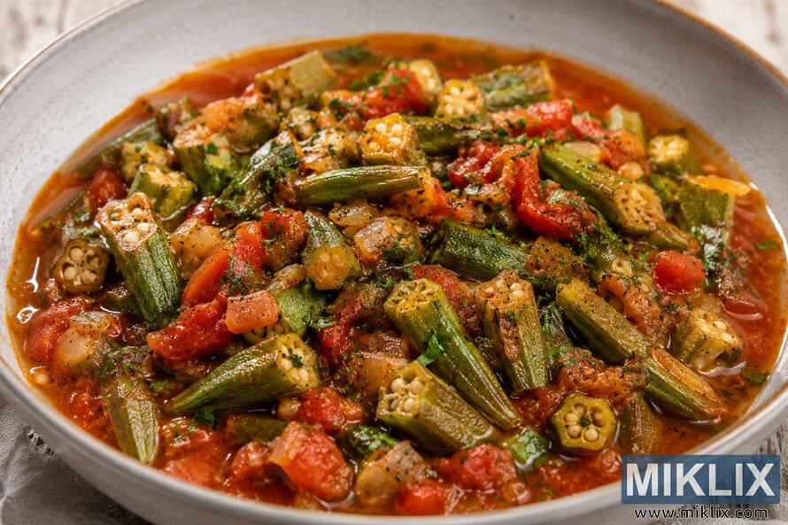 Close-up landscape photograph of a Mediterranean-style okra and tomato stew served in a rustic ceramic bowl, featuring tender okra, chunky tomatoes, onions, herbs, and a rich tomato broth on a weathered wooden surface under soft natural light. Close-up landscape photograph of a Mediterranean-style okra and tomato stew served in a rustic ceramic bowl, featuring tender okra, chunky tomatoes, onions, herbs, and a rich tomato broth on a weathered wooden surface under soft natural light.