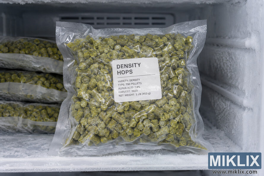 Vacuum-sealed bag of green Density hop pellets stored upright in a heavily frosted freezer, with additional sealed hop bags stacked in the background.
