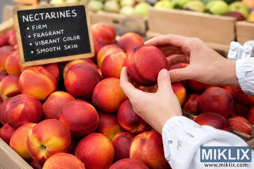 Close-up landscape photo of hands selecting ripe nectarines from wooden crates at a farmers market, with colorful fresh fruit and a chalkboard sign showing nectarine selection tips in the background. Close-up landscape photo of hands selecting ripe nectarines from wooden crates at a farmers market, with colorful fresh fruit and a chalkboard sign showing nectarine selection tips in the background.