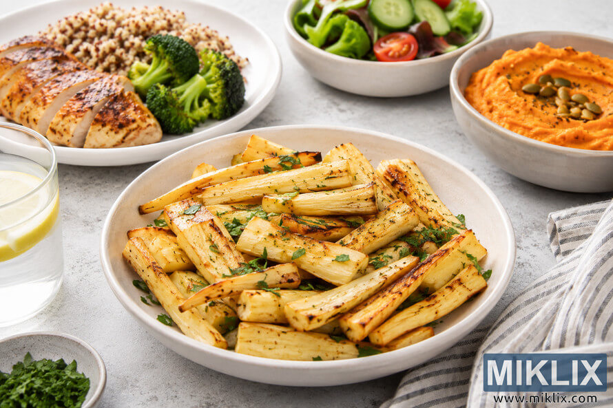 A landscape food photograph featuring a bowl of roasted parsnip batons surrounded by balanced meal components including grilled chicken, quinoa, broccoli, fresh salad, mashed sweet potato, herbs, and a glass of lemon water on a light stone surface. A landscape food photograph featuring a bowl of roasted parsnip batons surrounded by balanced meal components including grilled chicken, quinoa, broccoli, fresh salad, mashed sweet potato, herbs, and a glass of lemon water on a light stone surface.