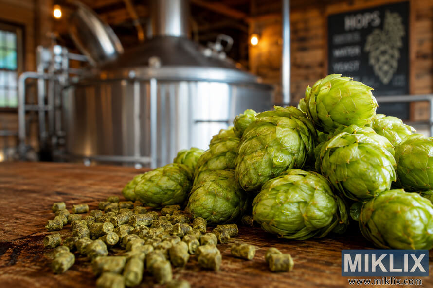Close-up of vibrant green Azacca hop cones with visible resin droplets and scattered hop pellets on a rustic wooden surface, with a softly blurred brewing kettle and warm brewery lighting in the background.