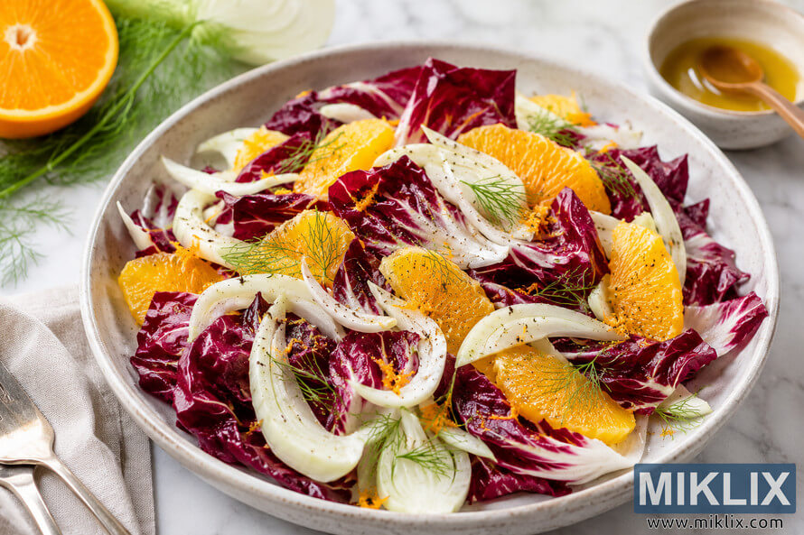 A vibrant citrus radicchio salad served in a rustic ceramic bowl, featuring deep purple radicchio leaves, bright orange segments, thinly sliced fennel, fennel fronds, orange zest, and cracked black pepper on a light marble countertop with simple table styling. A vibrant citrus radicchio salad served in a rustic ceramic bowl, featuring deep purple radicchio leaves, bright orange segments, thinly sliced fennel, fennel fronds, orange zest, and cracked black pepper on a light marble countertop with simple table styling.