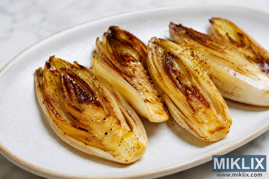 Close-up landscape photograph of grilled Belgian endive halves arranged on a white ceramic serving plate. The endive leaves are golden brown with deeply caramelized edges and light char marks, glistening with oil and seasoning under soft natural lighting on a pale marble surface. Close-up landscape photograph of grilled Belgian endive halves arranged on a white ceramic serving plate. The endive leaves are golden brown with deeply caramelized edges and light char marks, glistening with oil and seasoning under soft natural lighting on a pale marble surface.