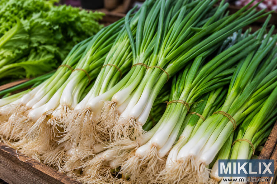 Bundles of fresh green scallions with white bulbs and long green stalks displayed on a rustic wooden crate at an outdoor farmers market, surrounded by leafy greens in soft natural light.