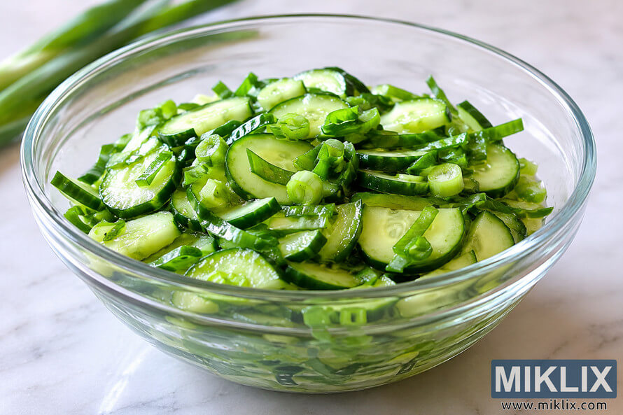 A fresh scallion and cucumber salad served in a transparent glass bowl on a light marble surface, featuring crisp cucumber slices and chopped green onions under soft natural lighting.