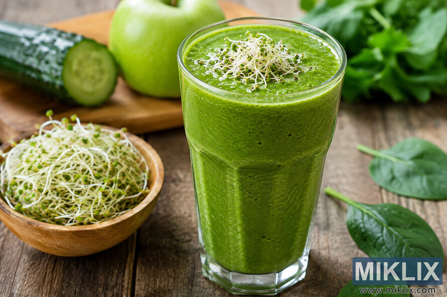 Fresh green smoothie in a tall glass topped with alfalfa sprouts, surrounded by cucumber, apple, spinach leaves, and a bowl of sprouts on a rustic wooden table.