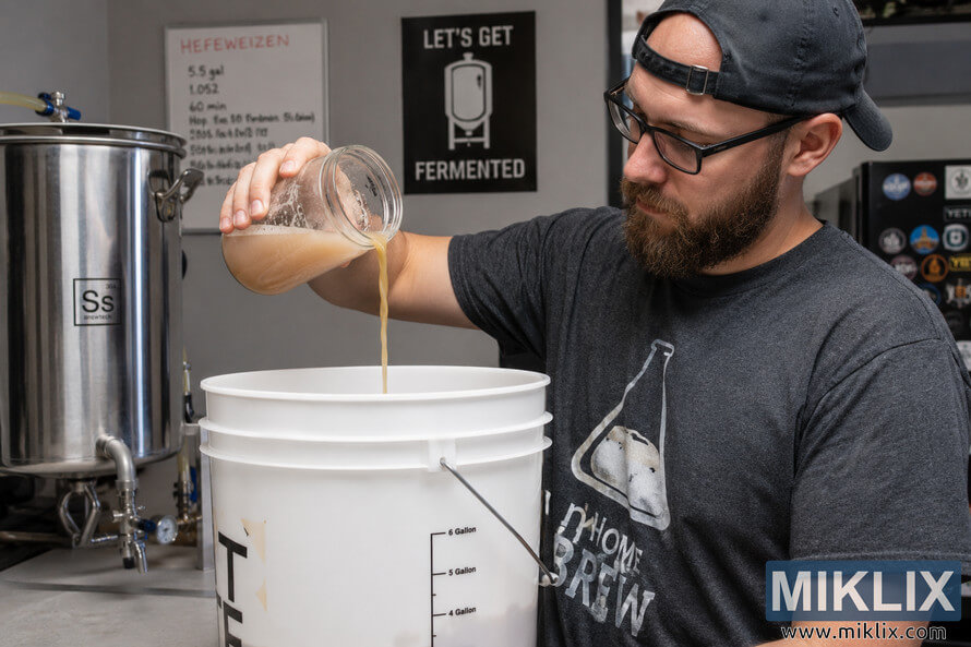 Homebrewer pouring a liquid yeast starter from a glass jar into a white fermentation bucket in a modern indoor brewing setup.