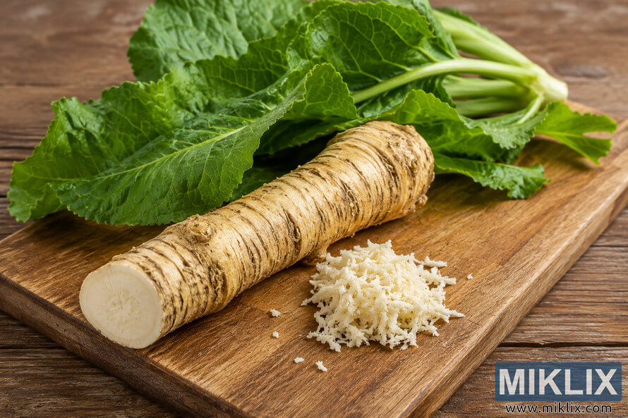 Fresh horseradish root with leafy green tops resting on a rustic wooden cutting board beside a small pile of grated horseradish in a warm kitchen setting.