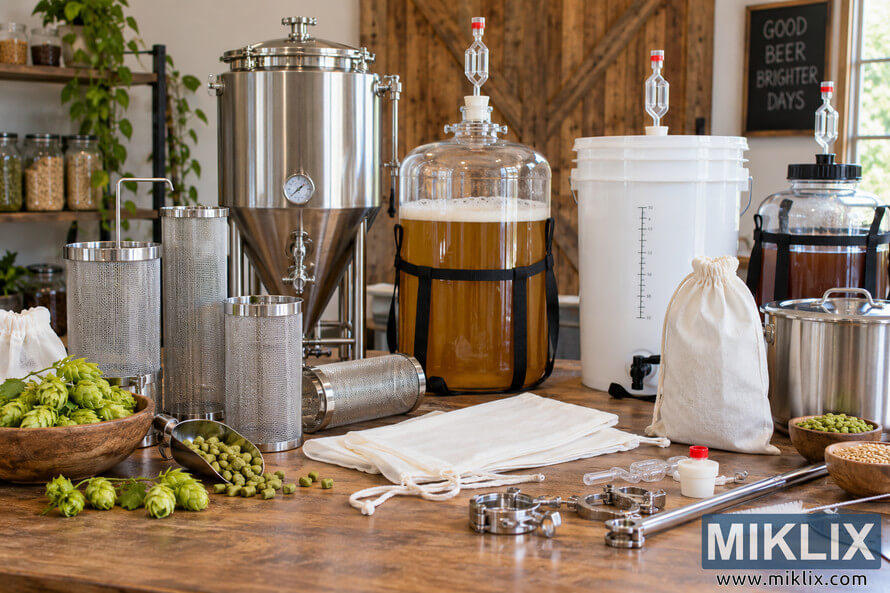 Landscape photo of a homebrew workspace with stainless-steel hop spiders, fabric brewing bags, fresh hops, hop pellets, grain, brewing fittings, a metal brewing vessel, and several glass and plastic fermenter options with airlocks. Landscape photo of a homebrew workspace with stainless-steel hop spiders, fabric brewing bags, fresh hops, hop pellets, grain, brewing fittings, a metal brewing vessel, and several glass and plastic fermenter options with airlocks.