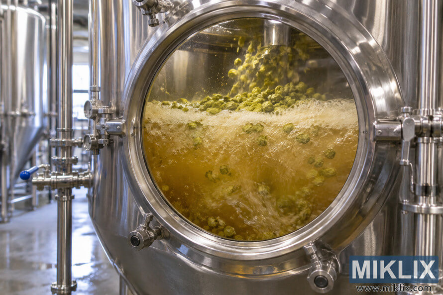 Close-up of a stainless-steel brewery fermenter with a circular viewing window showing golden beer actively fermenting with foam, bubbles, and green dry hop pellets swirling through the liquid. Close-up of a stainless-steel brewery fermenter with a circular viewing window showing golden beer actively fermenting with foam, bubbles, and green dry hop pellets swirling through the liquid.