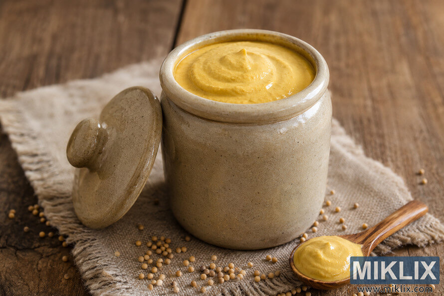 A rustic stoneware jar filled with creamy Dijon mustard sits on a weathered wooden table beside scattered mustard seeds and a wooden spoon with mustard, illuminated by soft natural light. A rustic stoneware jar filled with creamy Dijon mustard sits on a weathered wooden table beside scattered mustard seeds and a wooden spoon with mustard, illuminated by soft natural light.