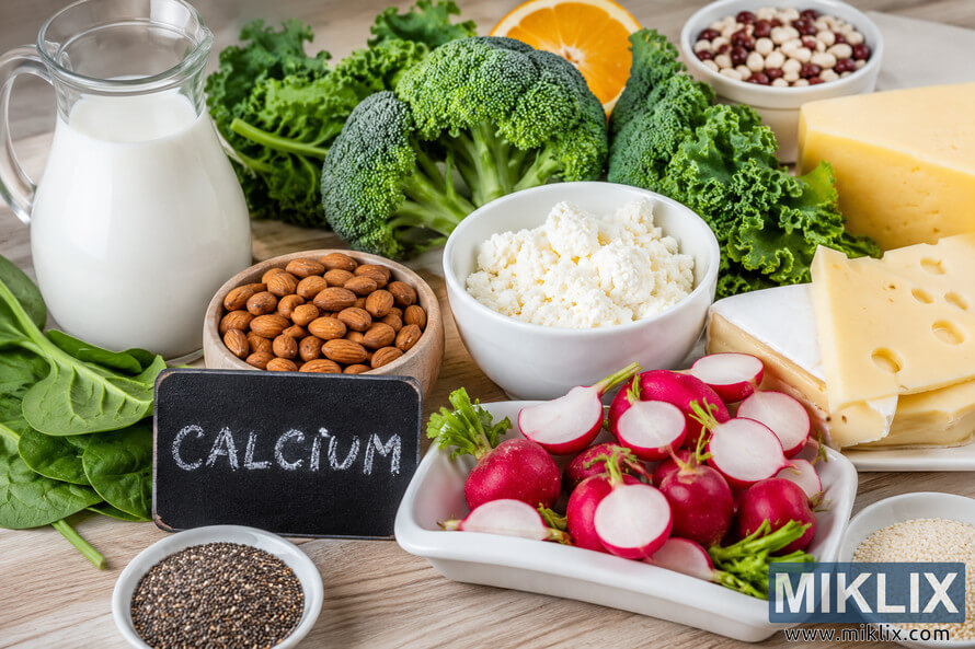 High-resolution landscape photograph of calcium-rich foods arranged on a wooden table, including milk, leafy greens, broccoli, cottage cheese, almonds, cheeses, chia seeds, sesame seeds, beans, a halved orange, and fresh whole and sliced radishes surrounding a small chalkboard labeled "CALCIUM," illustrating foods commonly associated with bone health.