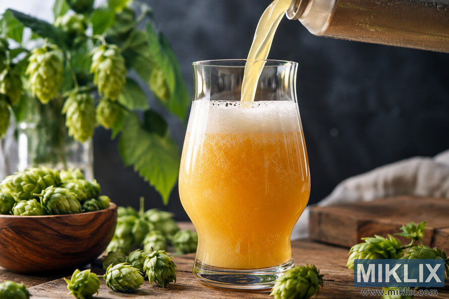 Hazy golden IPA being poured into a clear beer glass with a creamy white foam head, surrounded by fresh green hop cones on a rustic wooden table. Hazy golden IPA being poured into a clear beer glass with a creamy white foam head, surrounded by fresh green hop cones on a rustic wooden table.