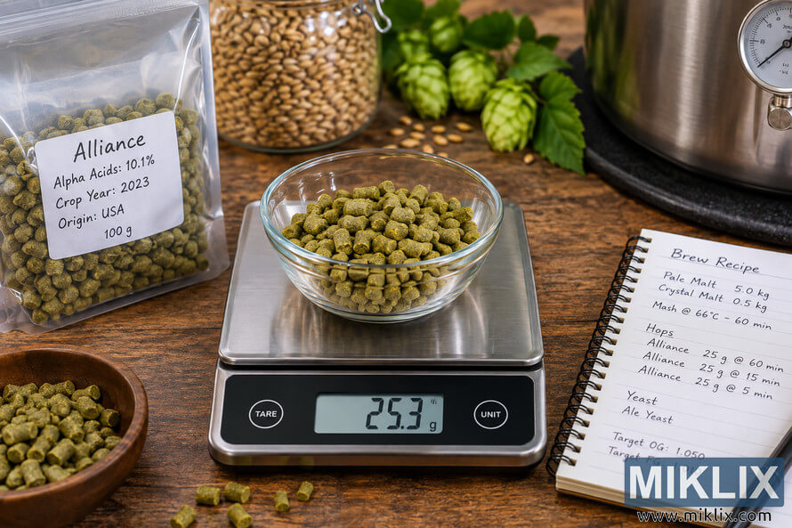 Landscape photo of a digital scale reading 25.3 g while weighing a glass bowl of green Alliance hop pellets on a wooden brewing table, with additional hops, malted grain, fresh hop cones, a handwritten beer recipe, and a stainless-steel brewing kettle in the background.