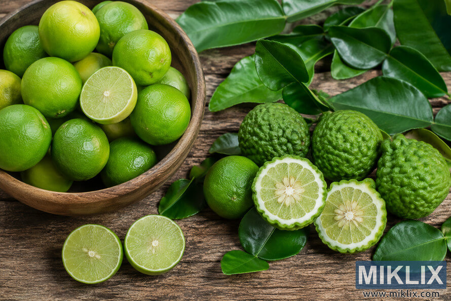 High-resolution landscape photograph of assorted green limes, including smooth key limes and textured makrut limes, arranged on a rustic wooden table with glossy citrus leaves and several halved fruits revealing their pale green interiors in natural daylight.