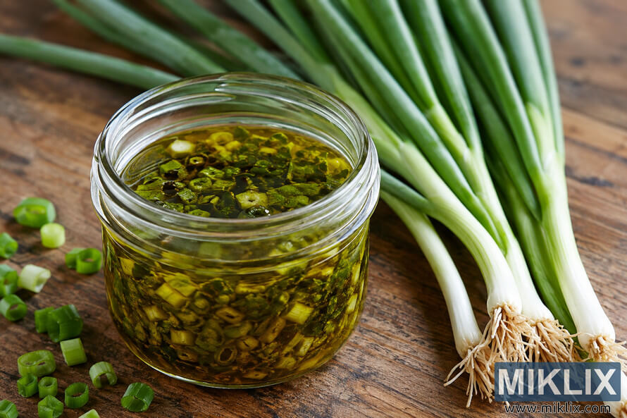 Close-up landscape photo of homemade scallion oil in a clear glass jar surrounded by fresh green onions on a rustic wooden surface, illuminated by soft natural light.