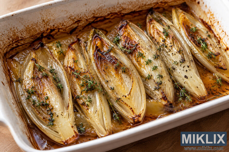 Close-up landscape photograph of braised Belgian endives arranged in a rustic ceramic baking dish, topped with herbs and featuring golden caramelized edges in warm natural light. Close-up landscape photograph of braised Belgian endives arranged in a rustic ceramic baking dish, topped with herbs and featuring golden caramelized edges in warm natural light.