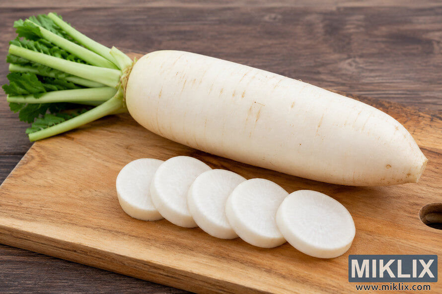 A whole white daikon radish with green leafy stems rests diagonally on a wooden cutting board beside five evenly sliced round pieces, photographed in a rustic kitchen with a warm wooden background.