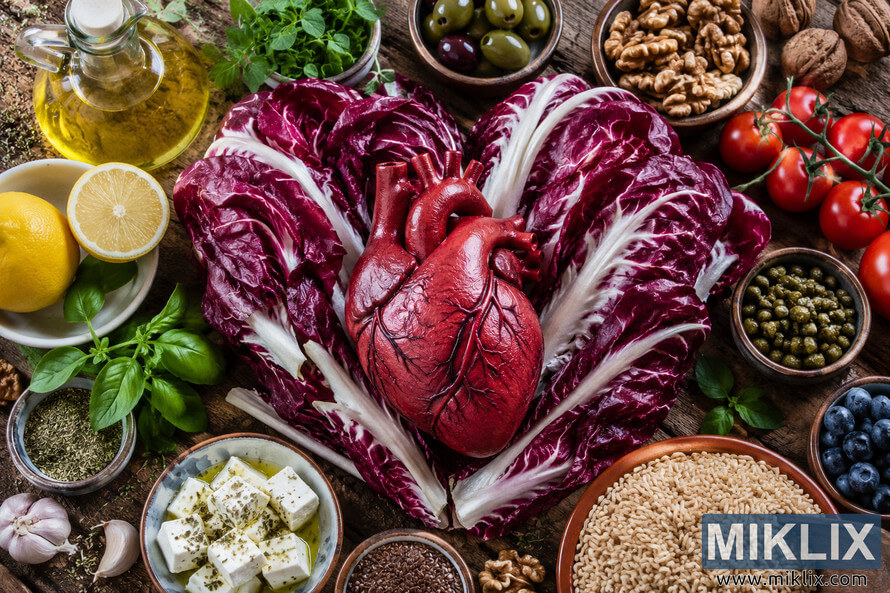 Top-down view of a realistic anatomical heart model resting on deep red radicchio leaves, surrounded by Mediterranean diet foods including olive oil, tomatoes, olives, walnuts, whole grains, herbs, blueberries, lemon, garlic, capers, flaxseed, and feta cheese on a rustic wooden table. Top-down view of a realistic anatomical heart model resting on deep red radicchio leaves, surrounded by Mediterranean diet foods including olive oil, tomatoes, olives, walnuts, whole grains, herbs, blueberries, lemon, garlic, capers, flaxseed, and feta cheese on a rustic wooden table.
