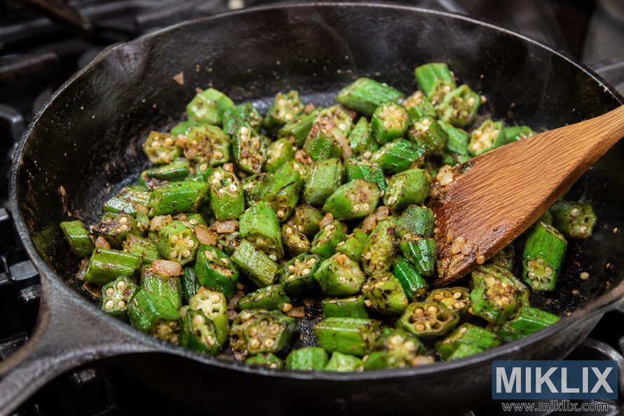 Close-up landscape view of freshly cut okra sautéing in a well-seasoned cast iron skillet on a stovetop while a wooden spatula stirs the vegetables as they caramelize and release steam. Close-up landscape view of freshly cut okra sautéing in a well-seasoned cast iron skillet on a stovetop while a wooden spatula stirs the vegetables as they caramelize and release steam.