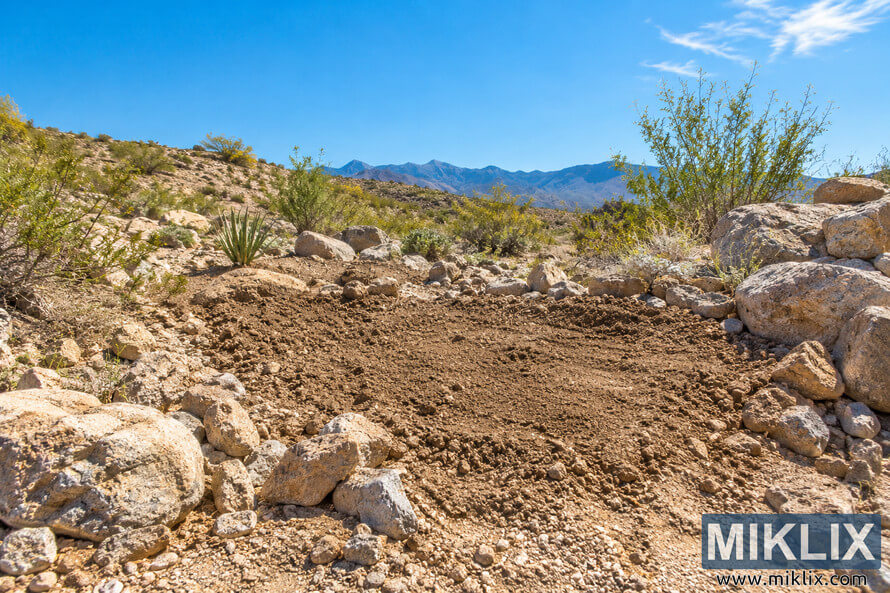 Sunny rocky desert planting site with freshly loosened soil, desert shrubs, boulders, and distant mountains. Sunny rocky desert planting site with freshly loosened soil, desert shrubs, boulders, and distant mountains.