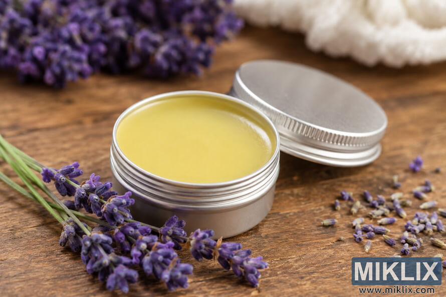 Close-up landscape image of a lavender healing balm in a silver metal tin resting on a rustic wooden surface, surrounded by fresh lavender sprigs and scattered dried lavender buds in warm natural lighting. Close-up landscape image of a lavender healing balm in a silver metal tin resting on a rustic wooden surface, surrounded by fresh lavender sprigs and scattered dried lavender buds in warm natural lighting.