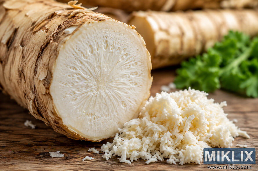 Close-up landscape photograph of a freshly cut horseradish root resting on a rustic wooden surface, showing its creamy white interior and fibrous texture alongside grated horseradish and soft green herbs in the blurred background.