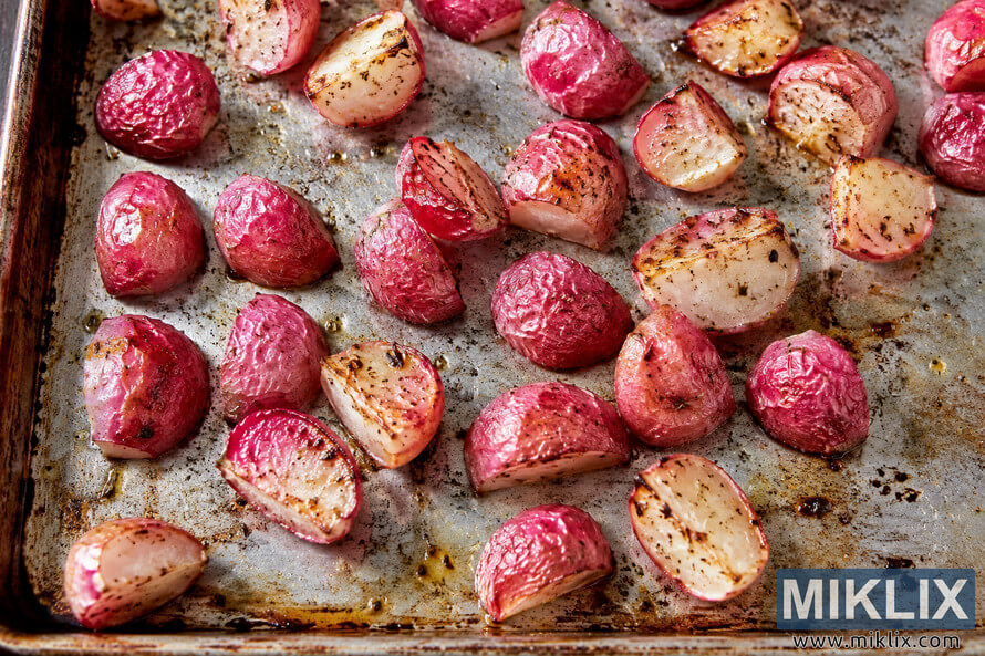 Overhead close-up of roasted pink radishes with caramelized edges, herbs, and oil scattered across a well-seasoned metal baking sheet.