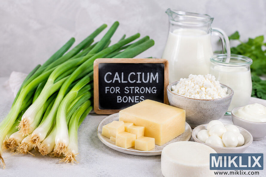 Fresh green scallions arranged beside assorted dairy products including milk, yogurt, cheese, cottage cheese, and mozzarella on a light neutral background with a small chalkboard sign reading 'Calcium for Strong Bones', symbolizing healthy calcium-rich foods that support bone health. Fresh green scallions arranged beside assorted dairy products including milk, yogurt, cheese, cottage cheese, and mozzarella on a light neutral background with a small chalkboard sign reading 'Calcium for Strong Bones', symbolizing healthy calcium-rich foods that support bone health.