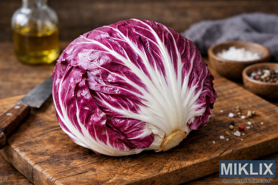 A fresh head of radicchio with vibrant purple leaves and white veins resting on a rustic wooden cutting board, surrounded by a knife, olive oil bottle, and small bowls of salt and peppercorns. A fresh head of radicchio with vibrant purple leaves and white veins resting on a rustic wooden cutting board, surrounded by a knife, olive oil bottle, and small bowls of salt and peppercorns.