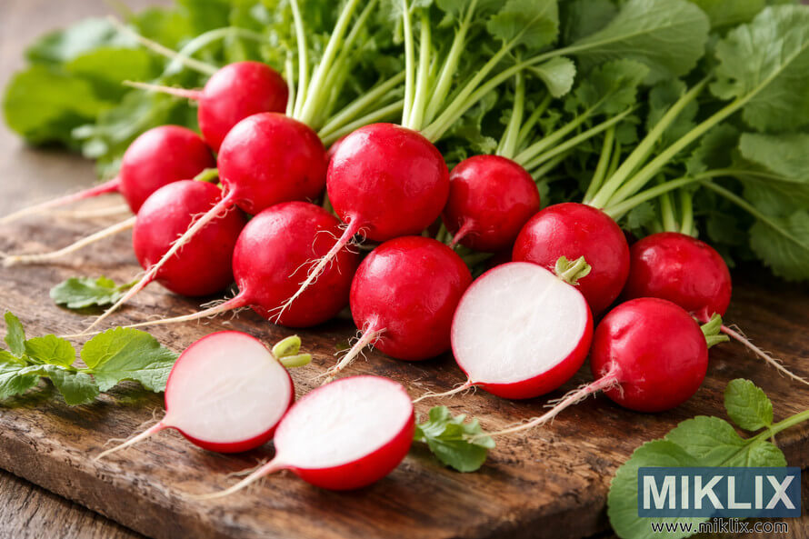 Round red Cherry Belle radishes with green tops, some sliced to reveal their crisp white flesh on a rustic wooden surface