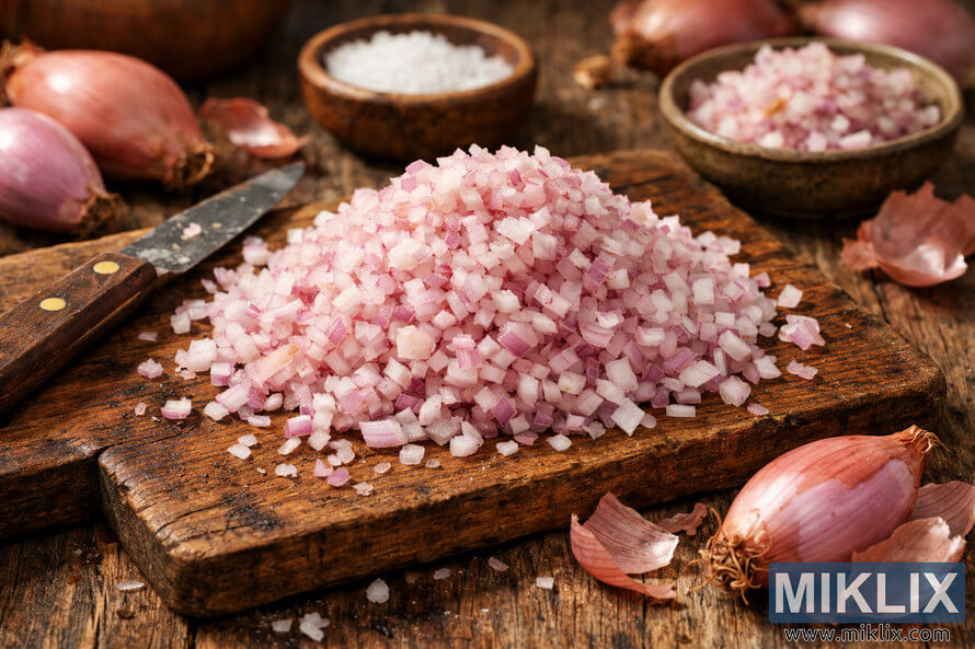 Finely minced shallots displayed on a rustic wooden table with whole shallots, knife, and ceramic dish in warm natural light.