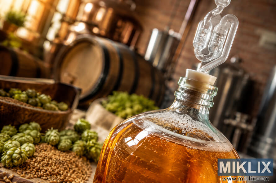 Close-up of a glass fermentation vessel filled with golden ESB-style ale during active fermentation, featuring a bubbling airlock, scattered hops and malt grains on a rustic wooden surface, with softly blurred brewery equipment and wooden barrels illuminated by warm natural light. Close-up of a glass fermentation vessel filled with golden ESB-style ale during active fermentation, featuring a bubbling airlock, scattered hops and malt grains on a rustic wooden surface, with softly blurred brewery equipment and wooden barrels illuminated by warm natural light.