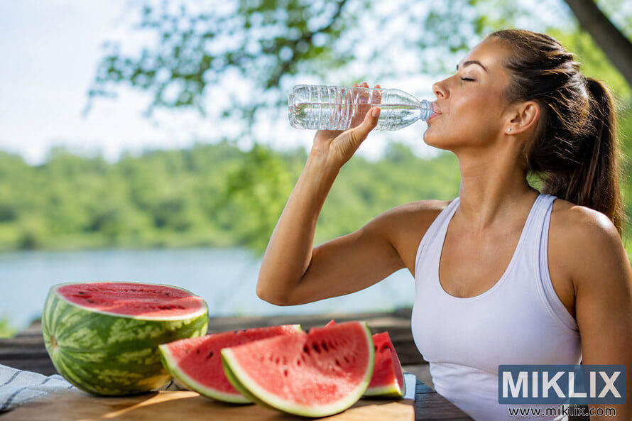 A person drinks water from a clear unbranded bottle beside freshly sliced watermelon on a wooden table in a sunny outdoor setting with trees and a lake in the softly blurred background, illustrating healthy hydration during summer. A person drinks water from a clear unbranded bottle beside freshly sliced watermelon on a wooden table in a sunny outdoor setting with trees and a lake in the softly blurred background, illustrating healthy hydration during summer.