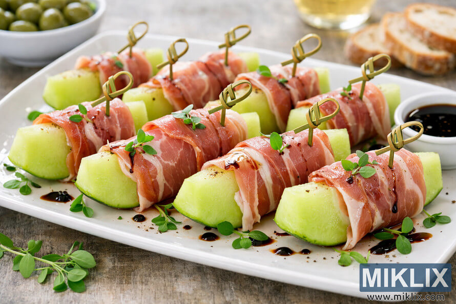 A platter of prosciutto-wrapped honeydew melon appetizers garnished with microgreens and balsamic glaze. A platter of prosciutto-wrapped honeydew melon appetizers garnished with microgreens and balsamic glaze.