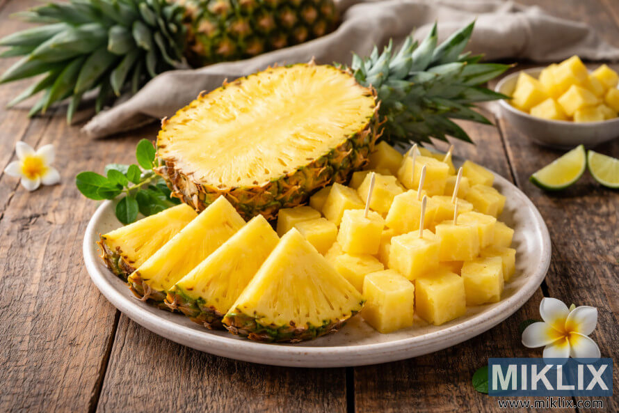 Pinya tallada per la meitat amb falques i daus en un plat beix, colÂ·locat sobre una taula de fusta rÃºstica amb detalls tropicals al fons.