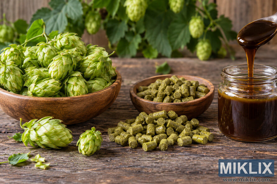 Rustic still life showing fresh green whole hop cones, compressed hop pellets, and dark amber hop extract in a glass jar on a weathered wooden table.