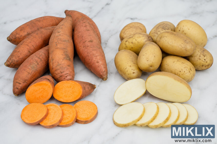 Landscape photo showing sweet potatoes on the left and white potatoes on the right arranged side by side on a light marble countertop, with both whole potatoes and sliced cross-sections highlighting the bright orange flesh of sweet potatoes and the creamy white flesh of regular potatoes. Landscape photo showing sweet potatoes on the left and white potatoes on the right arranged side by side on a light marble countertop, with both whole potatoes and sliced cross-sections highlighting the bright orange flesh of sweet potatoes and the creamy white flesh of regular potatoes.