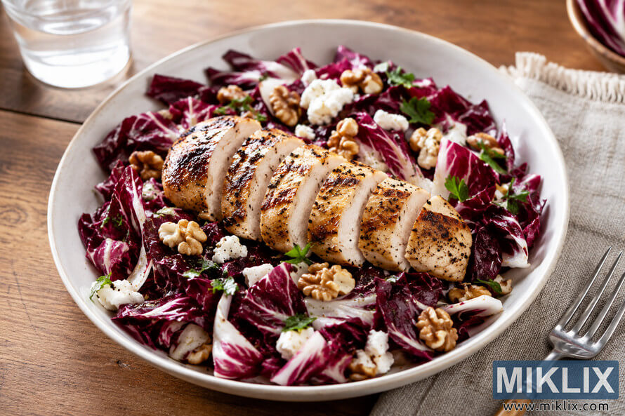 Landscape food photograph of sliced grilled chicken breast served over a fresh radicchio salad with walnuts, crumbled white cheese, herbs, and light dressing in a ceramic bowl on a rustic wooden table, accompanied by a glass of water, napkin, and fork in soft natural light. Landscape food photograph of sliced grilled chicken breast served over a fresh radicchio salad with walnuts, crumbled white cheese, herbs, and light dressing in a ceramic bowl on a rustic wooden table, accompanied by a glass of water, napkin, and fork in soft natural light.