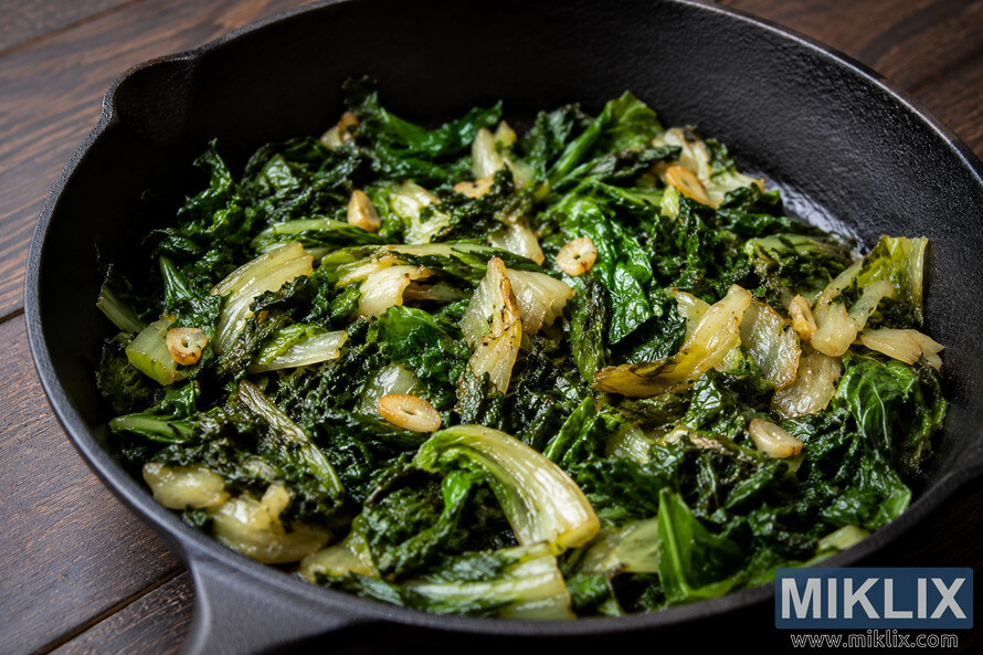 Close-up of sautéed escarole with sliced garlic cooking in a black cast iron skillet on a rustic wooden table. The greens are glossy and tender with lightly caramelized edges under warm natural lighting. Close-up of sautéed escarole with sliced garlic cooking in a black cast iron skillet on a rustic wooden table. The greens are glossy and tender with lightly caramelized edges under warm natural lighting.