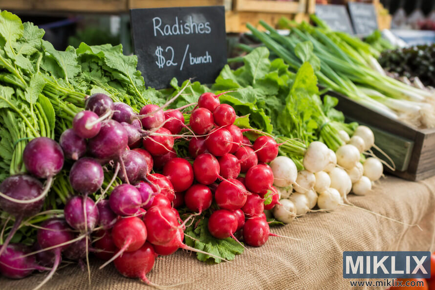 Landscape photograph of fresh red, purple, and white radishes with leafy green tops arranged on a burlap-covered farmers market table beside a handwritten chalkboard price sign, with softly blurred vegetables in the background.