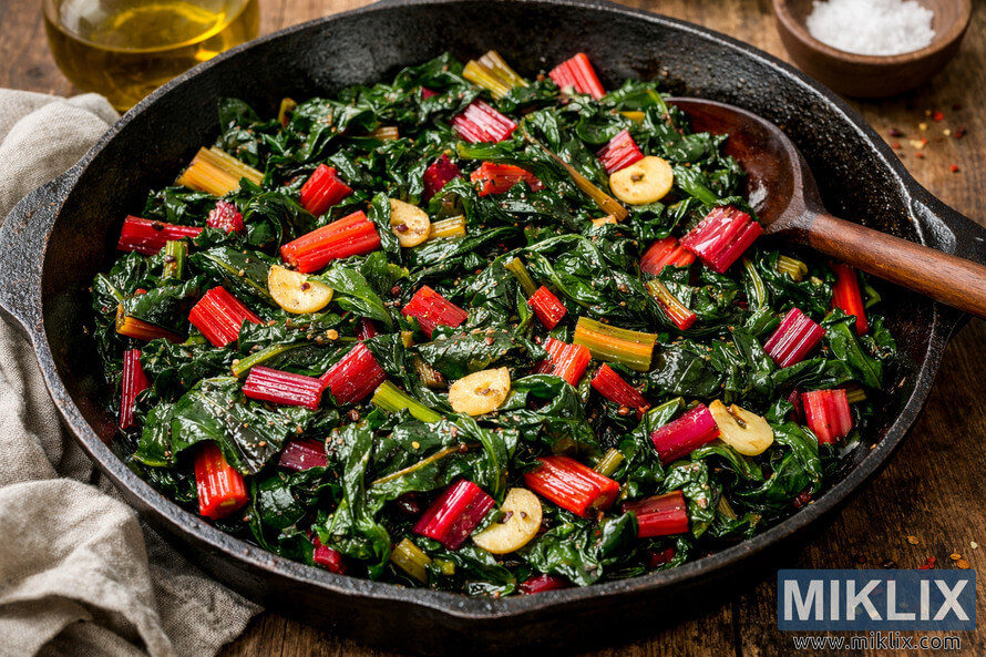 A close-up view of cooked Swiss chard in a rustic cast iron skillet, glistening with olive oil and golden garlic slices.