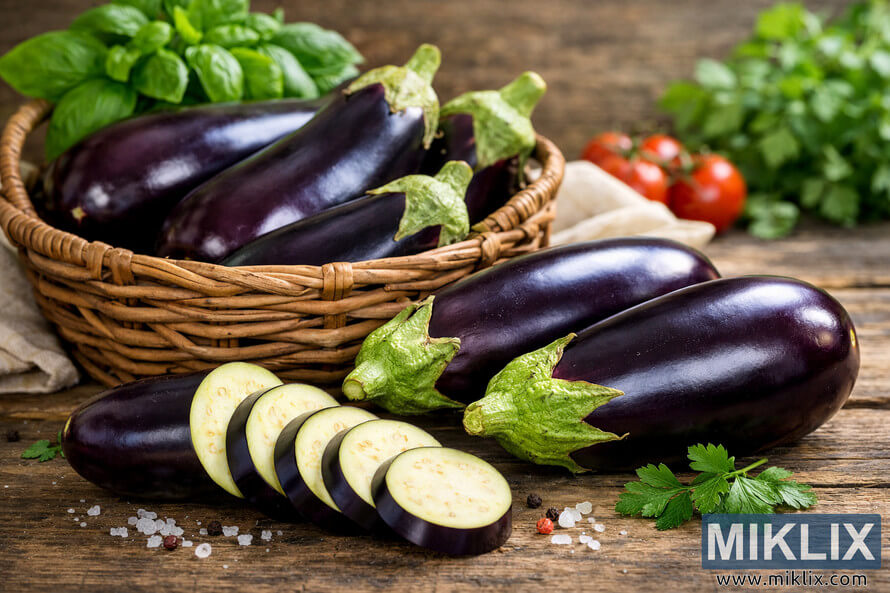 Freshly harvested eggplants in a wicker basket with sliced pieces on a rustic wooden table surrounded by herbs and spices. Freshly harvested eggplants in a wicker basket with sliced pieces on a rustic wooden table surrounded by herbs and spices.
